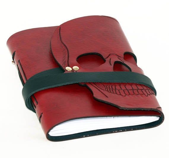 features dark red leather journal with skull on cover. there are two gold bolts near temple of skull to fasten a black leather strap which is used to close journal. bright white pages are seen at bottom of journal. white background