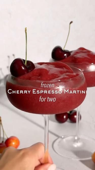 two drinks featuring a drink called a " frozen Cherry Espresso Martini". Hand in bottom left corner holding one of the drinks. Drinks feature a cherry in each glass. there are a few cherries on white table the drinks sit upon