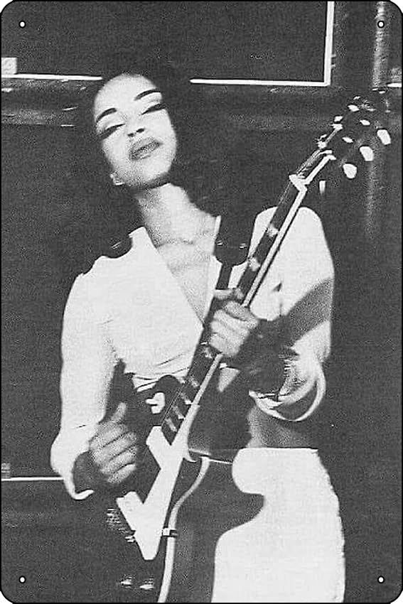 A black and white photo of musician Sade Adu. She has a serene facial expression while playing the guitar onstage. She wears a white two-piece outfit, long-sleeved cropped top and what appears to be a long skirt. 