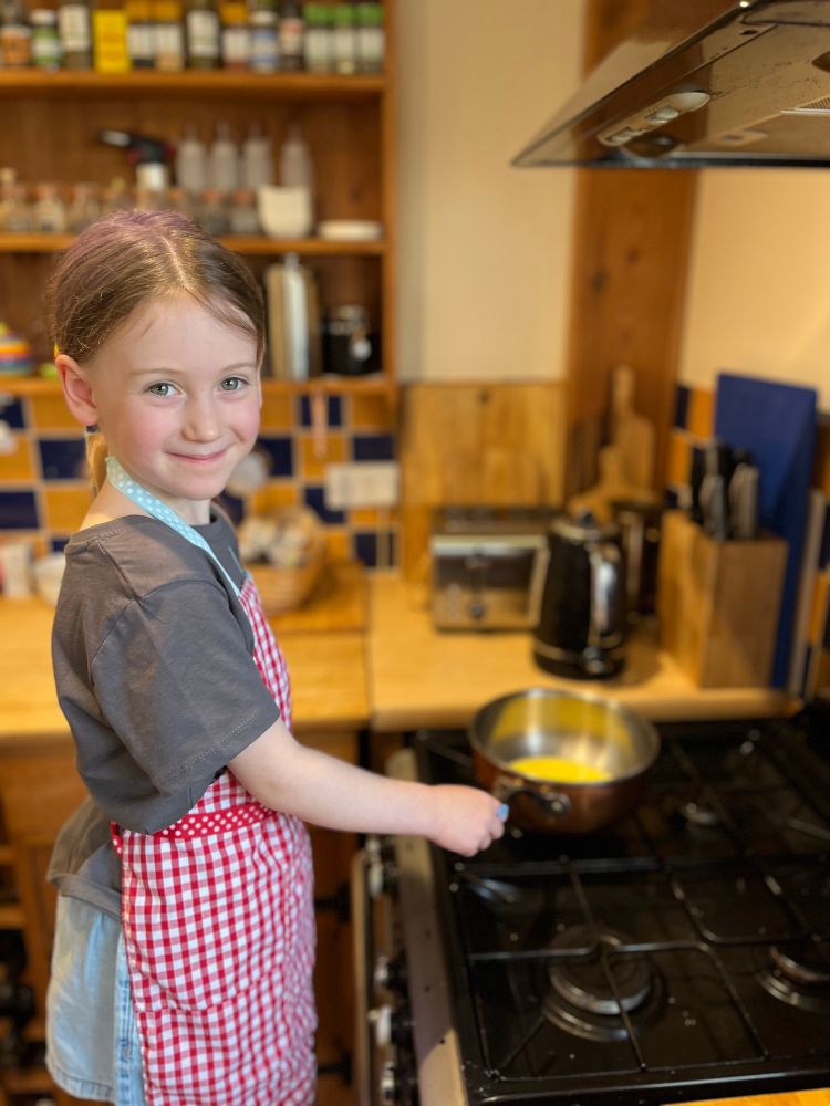 A young girl in a red chequered apron holding a copper saucier pan on a gas oven. There is milk and melted butter in the pan. 