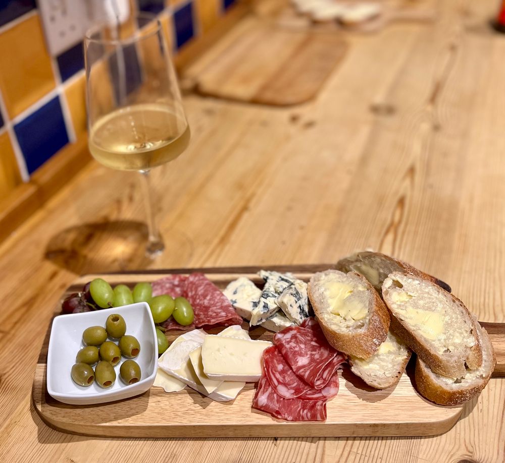 A cheeseboard with olives, Brie and blue cheeses, salamis and sliced baguette. A glass of white wine is behind it. 