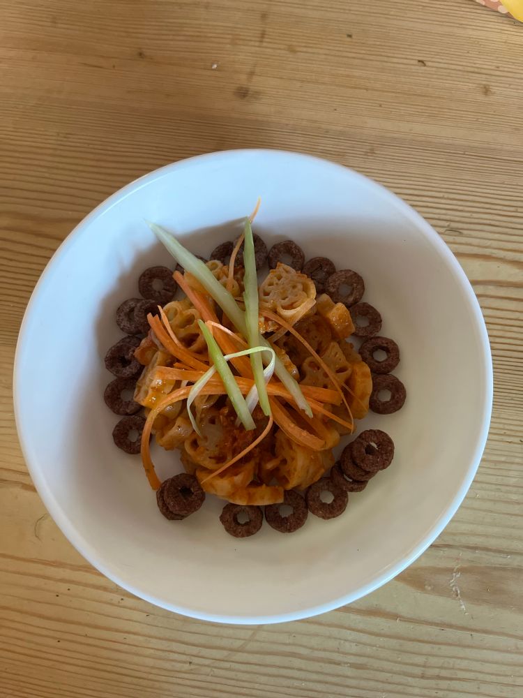A bowl containing Paw Patrol pasta shapes in red pesto, garnished with julienne carrots and celery, surrounded by Choco Hoops. 