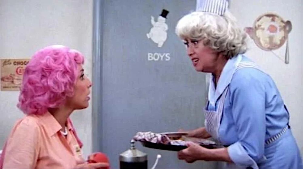 Didi Conn as 'Frenchie' with Joan Blondell as 'Vi' at the Frosty Palace diner in GREASE (1978). 'Frenchie' has bright pink hair as a result of a tinting mishap in beauty school. The restroom door has a heavy snowman wearing a top hat. 'Vi' carries a tray, empty except for a washcloth.