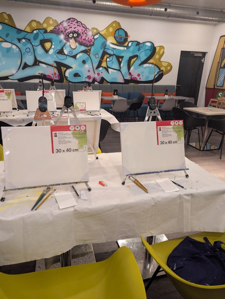 Ein länglicher Tisch mit Leinwänden und Mal-Utensilien, dazu bunte Stühle. Im Hintergrund noch ein Tisch. Graffiti an der Wand. 