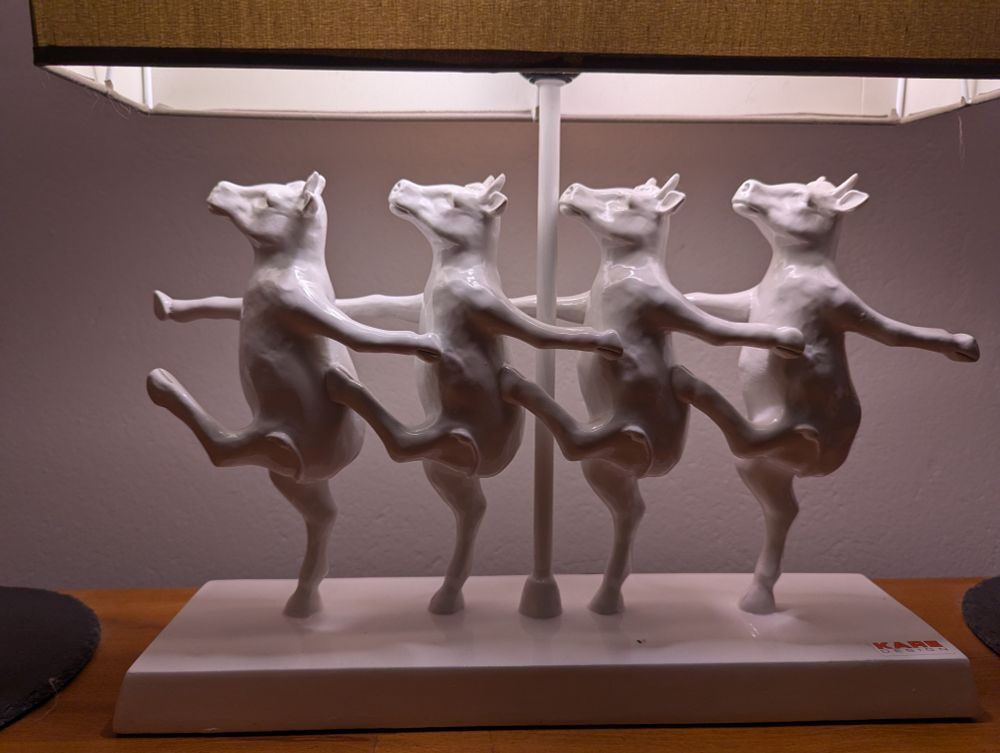 4 gleich tanzende weiße keramik-Kühe auf den Hingerbeinen wobei eines davon erhoben ist unter einem rechteckigen Lampenschirm 