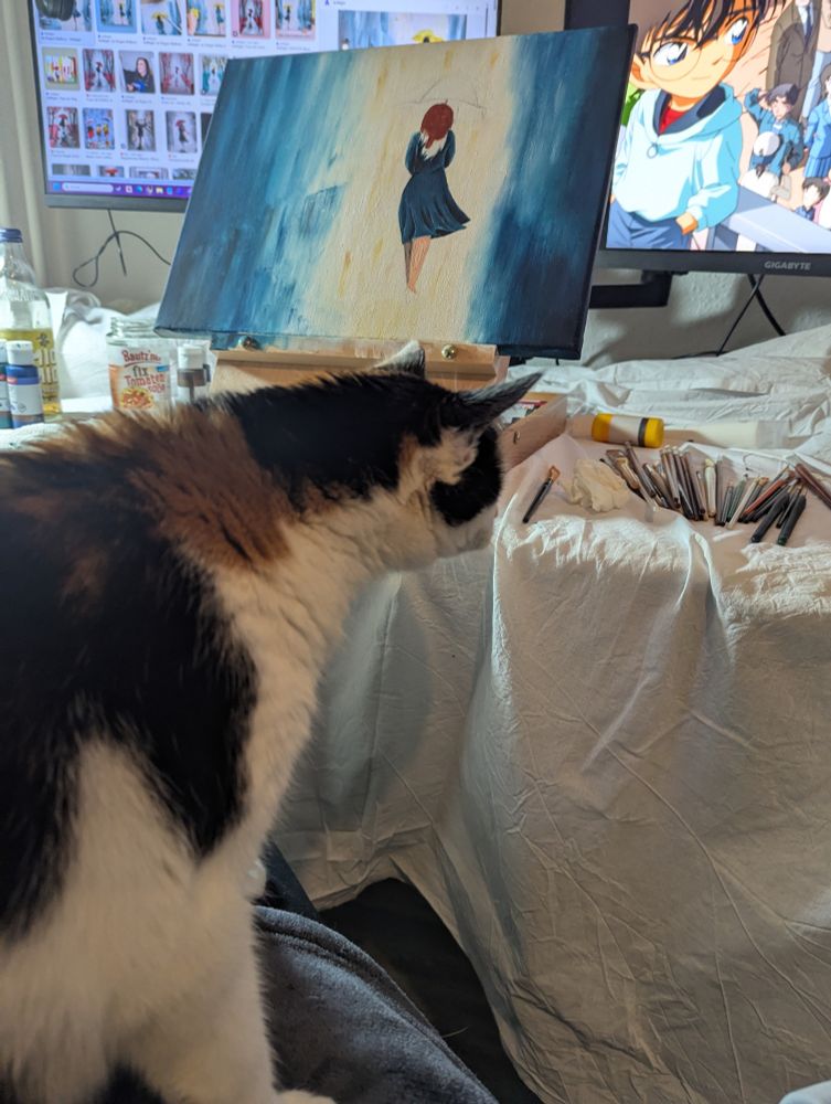 Tisch bedeckt mit einem weißen Bettlaken voller Malkram (Pinsel und Farben sowie ein Tischstaffelei mit Bild) dahinter Bildschirme. 
Eine Katze auf meinem Schoß macht langen Hals und schaut interessiert auf den Tisch 