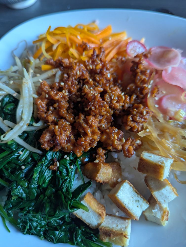 Bild eines Tellers mit Essen. Zu sehen sind Spinat, Sprossen, Karotten, Radieschen, Zwiebel und Tofu-würfel alles nacheinander im Kreis angeordnet. In der Mitte veganes Hack. 