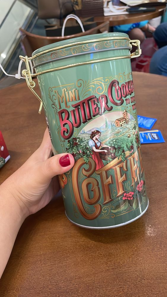 To segurando uma lata de biscoitos daquelas de vó. A lata é comprida e tem um design retrô com cores verde-água, vermelho e detalhes dourado. Tá escrito “butter cookies to enjoy with coffee” 

Essa maior custou R$ 40 nas Lojas americanas. 