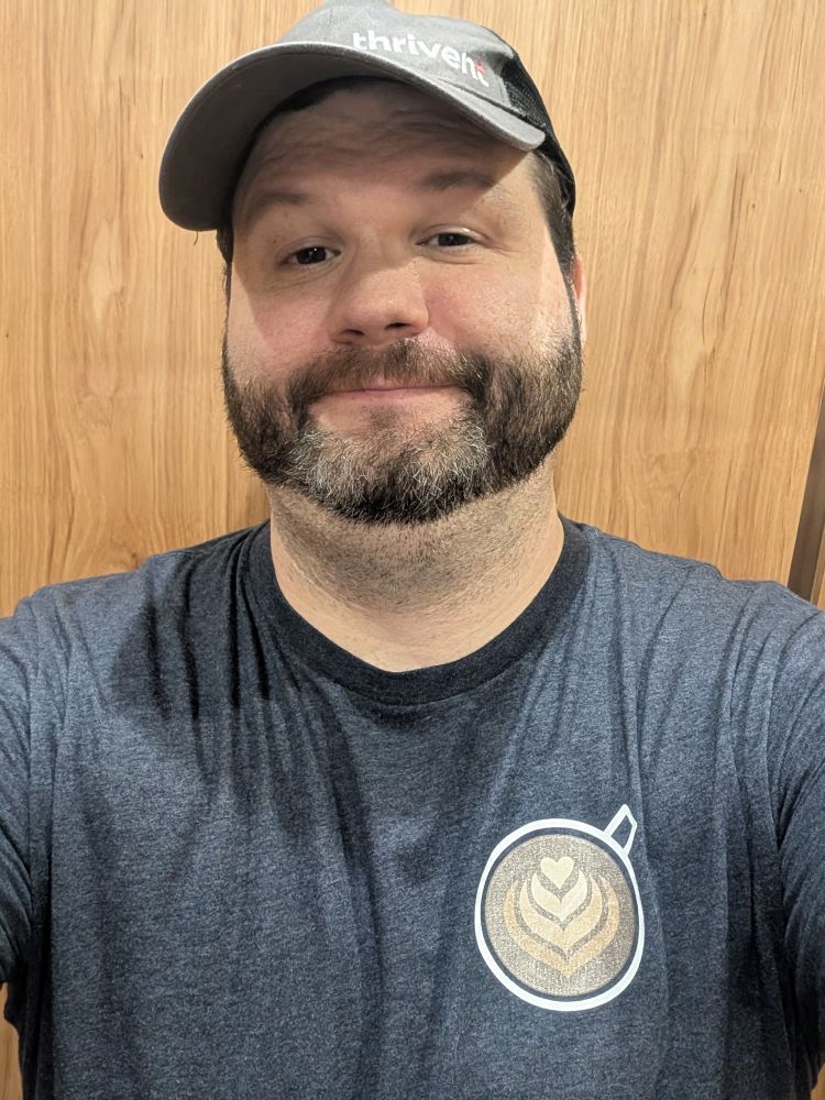 Me, a guy with a graying beard and sleepy dark eyes, posing for a selfie in front of a wood wall wearing a gray shirt with a picture of a top down latte on it, also wearing my gray work cap.