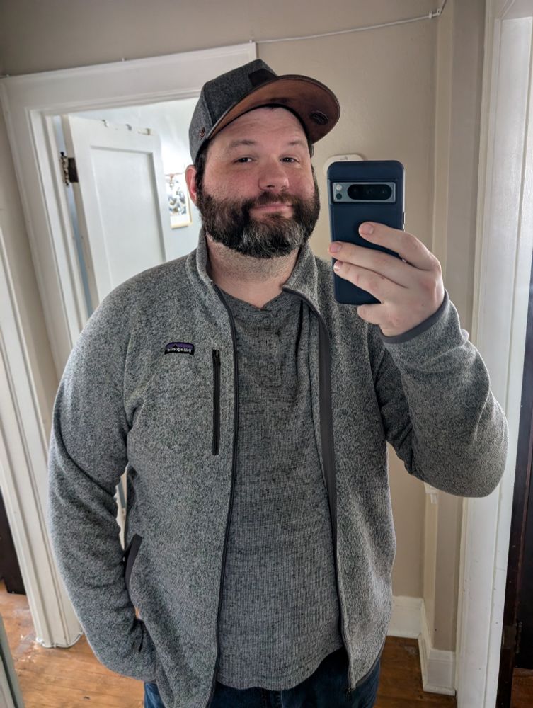 A selfie in the mirror of me, a big guy with a beard wearing a gray hat, gray shirt, gray jacket, and blue jeans.