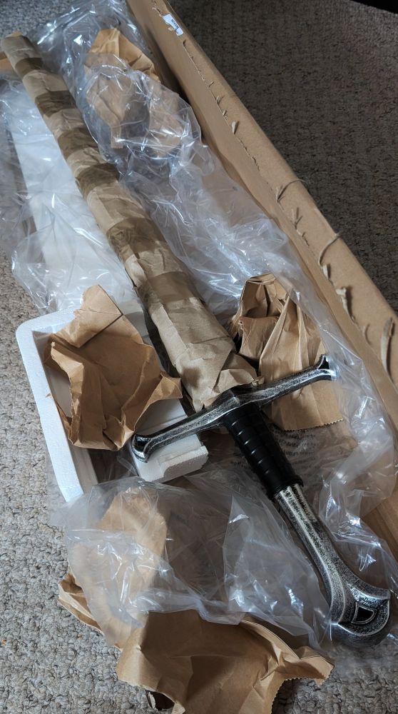 A large sword with a black handle partially wrapped in brown paper and plastic sits inside an open cardboard shipping box on a carpeted floor.