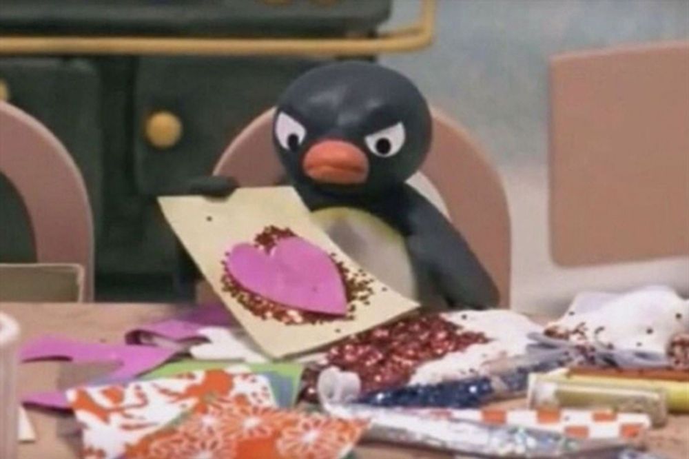 Pingu, der Pinguin aus der gleichnamigen Knetanimation-Trickfilmserie, sitzt mit grimmigem Gesichtsausdruck an einem Tisch und bastelt eine Karte. Die Karte ist mit funkelndem Glitzer verziert und zeigt ein großes, pinkes Herz in der Mitte. Der Pinguin wirkt konzentriert und entschlossen, während er an seinem Bastelprojekt arbeitet.