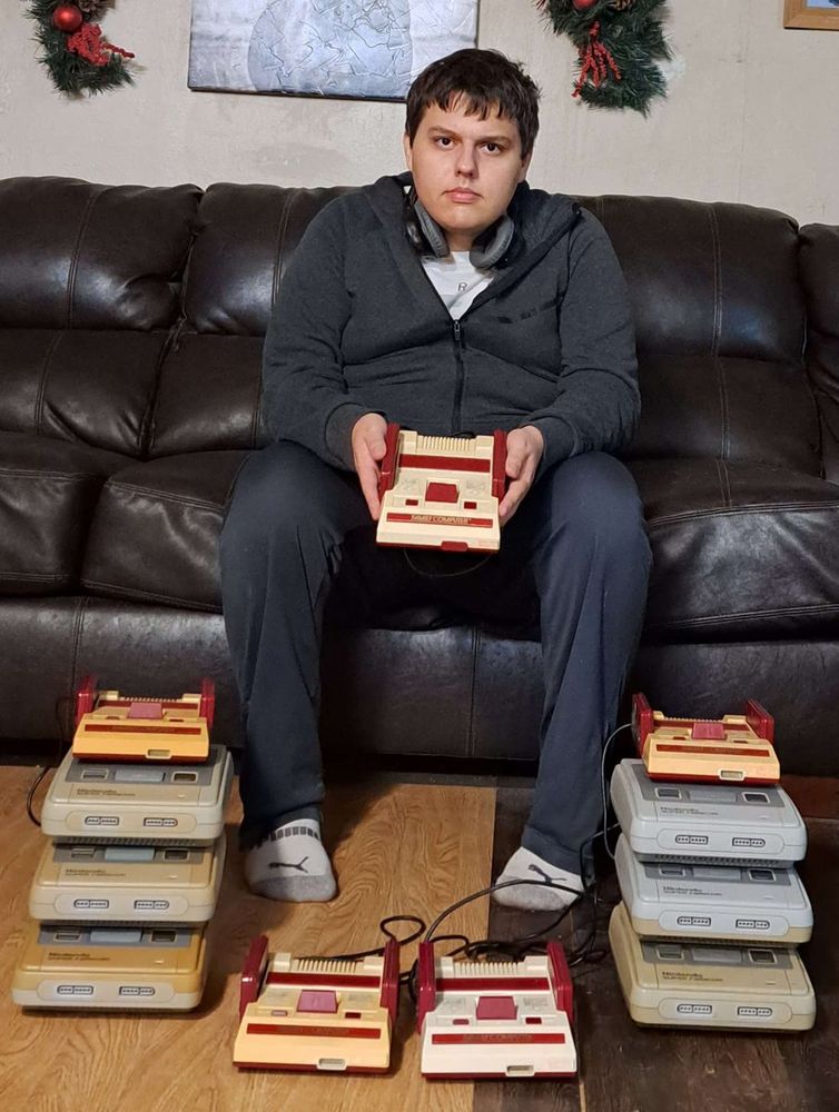 Photograph of Brenden holding a Famicom with two Famicom consoles on the floor as well as two stacks of three Super Famicoms with a Famicom on top of each stack.