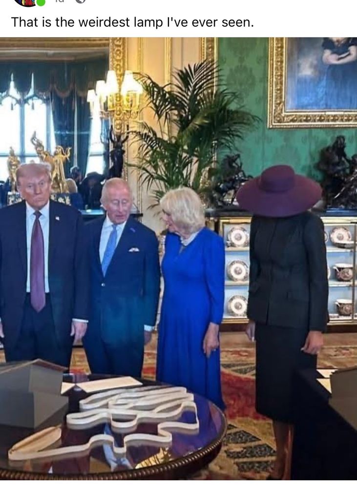An indoors picture of The Orange 🤡, King Charles, Camilla, and Mel who is wearing her dark purple hat such that it looks like a lampshade.