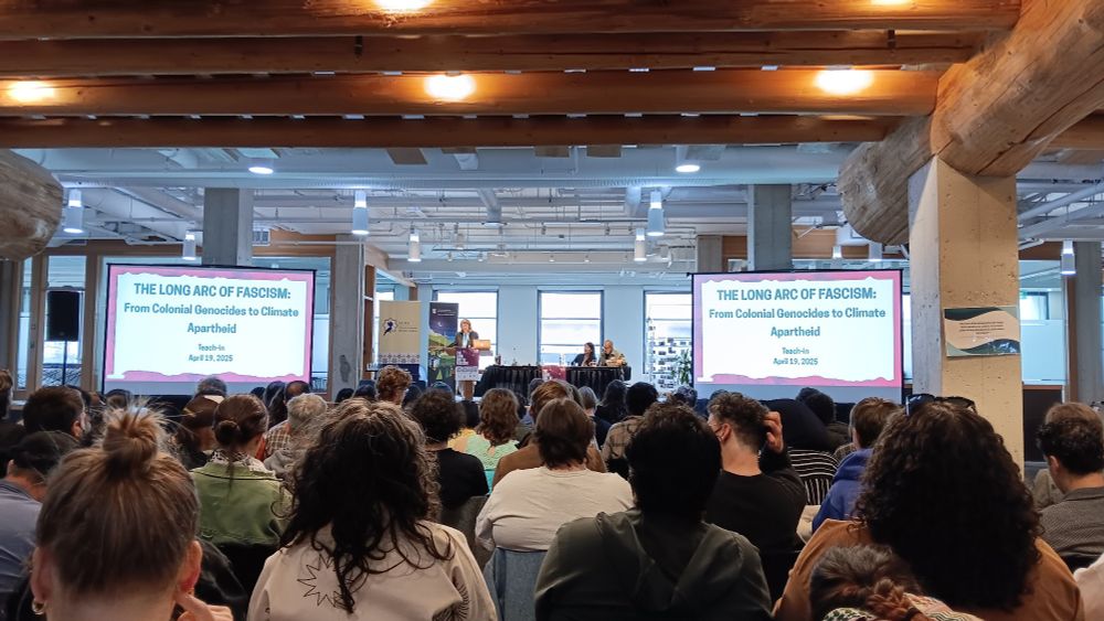 Wide picture shot showing many people seated listening to Naomi Klein speaking at a podium beside a projector slide saying The Long Arc of Fascism. 