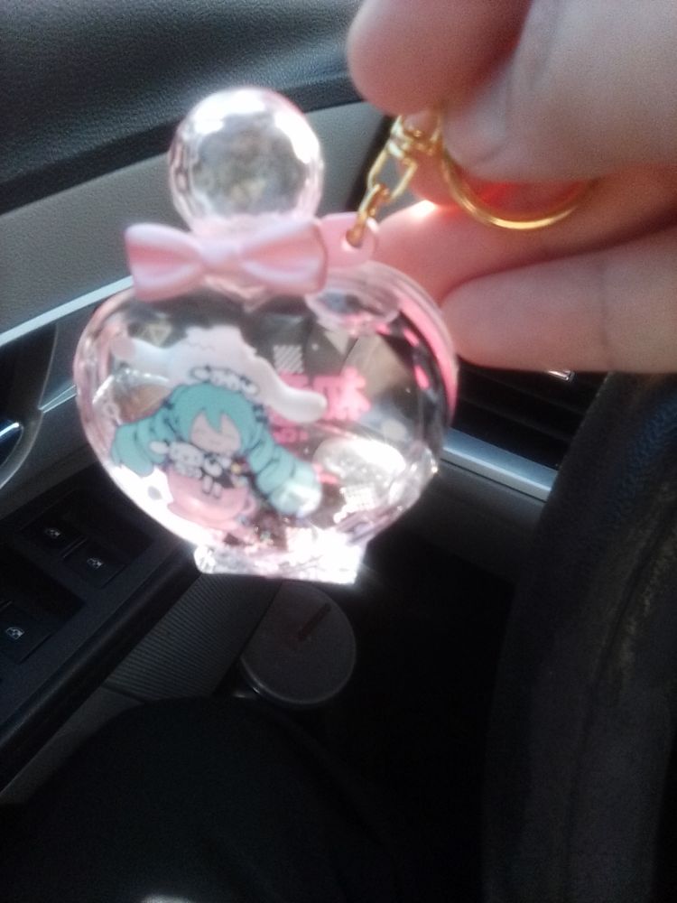 A pink bottle shaped keychain filled with water, gel, and a tiny cinnamaroll. The bottle has a picture of Miku in an almost maid style dress sitting on a tea cup with two little cinnamarolls