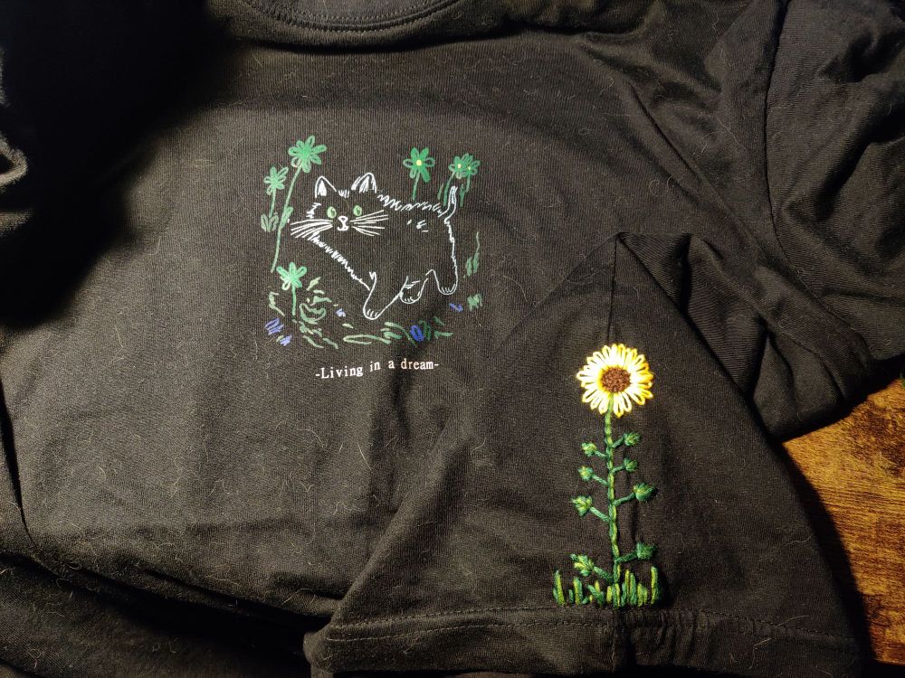 Shirt with a print of a cat in a flower field with "Living in a dream" written under it, on the side of the shirt is an embroidery of a sunflower 