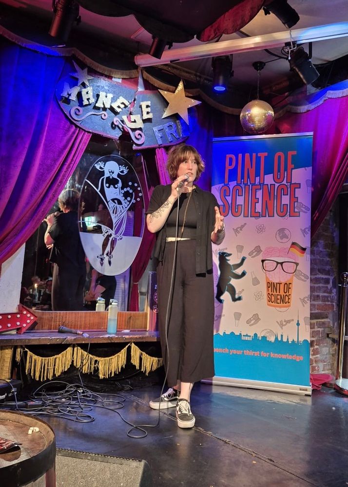 A person dressed in black in on a stage with a microphone. There is some burlesque imagery, and a Pint of Science banner.