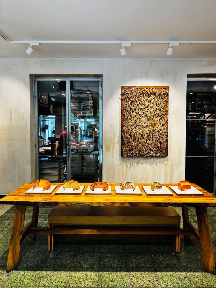 Ein Geschäft, in der Mitte ein Tisch auf dem 6 Brote liegen. Der Boden ist grün gefliest, die Wand weiß und es hängt ein Bild mit Getreidepflanzen an der Wand. Durch ein Fenster hat man Einblick in eine Backstube