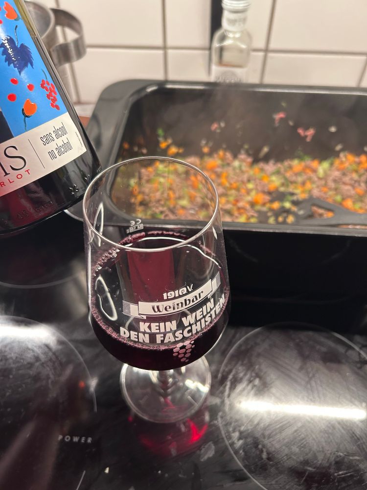 Ein Glas Rotwein auf einem Glaskeramikkochfeld. Auf dem Glas steht „kein Wein den Faschisten“ 
im Hintergrund köchelt Bolognese im Bräter.
Am linken Bildrand die Flasche Rotwein auf der „sans alcohol/no alcohol“ steht