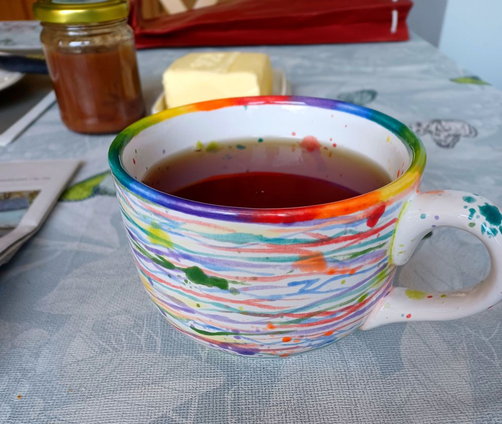 Eine Tasse mit vielen bunten Streifen und Kleksen und einem Regenbogenrand. Darin ist schwarzer Tee. 