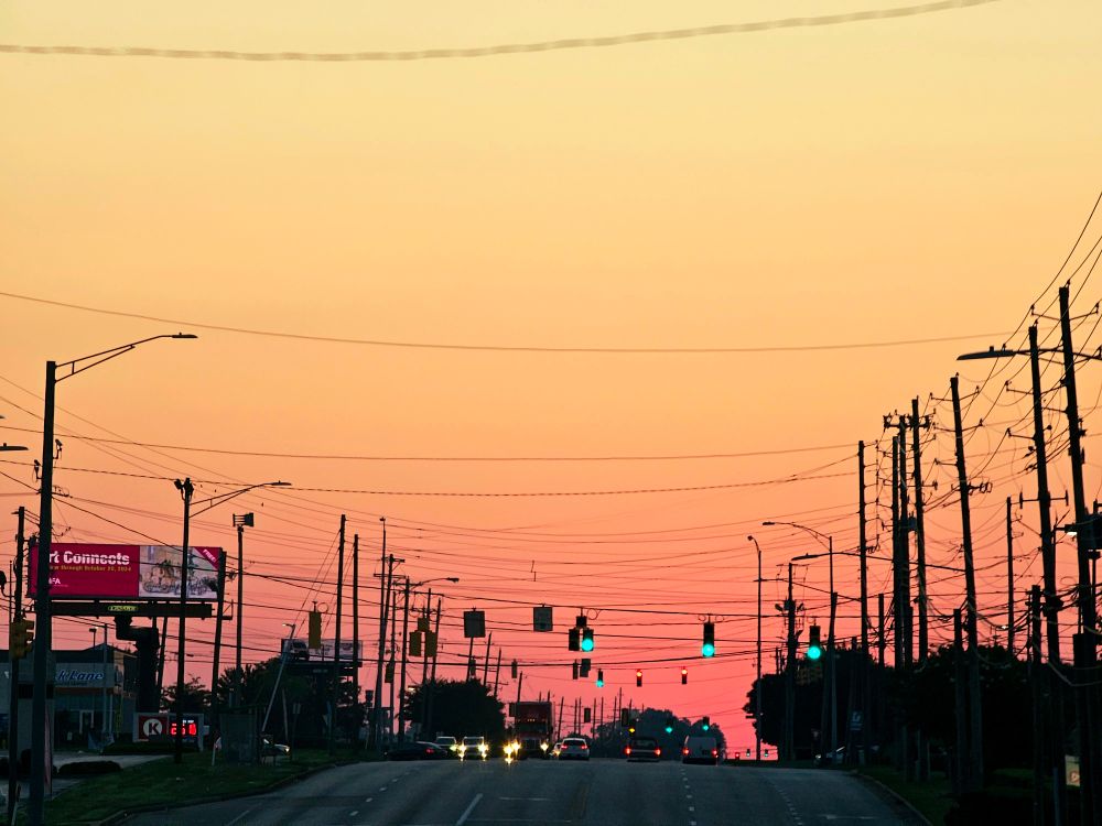 A morning street set to a sunrise background with a harsh contrast. Most of the foreground is a black silhouette, while the sky is a bright red-orange and yellow.
Traffic lights and signal lamps light the street below. There is one billboard to the left.