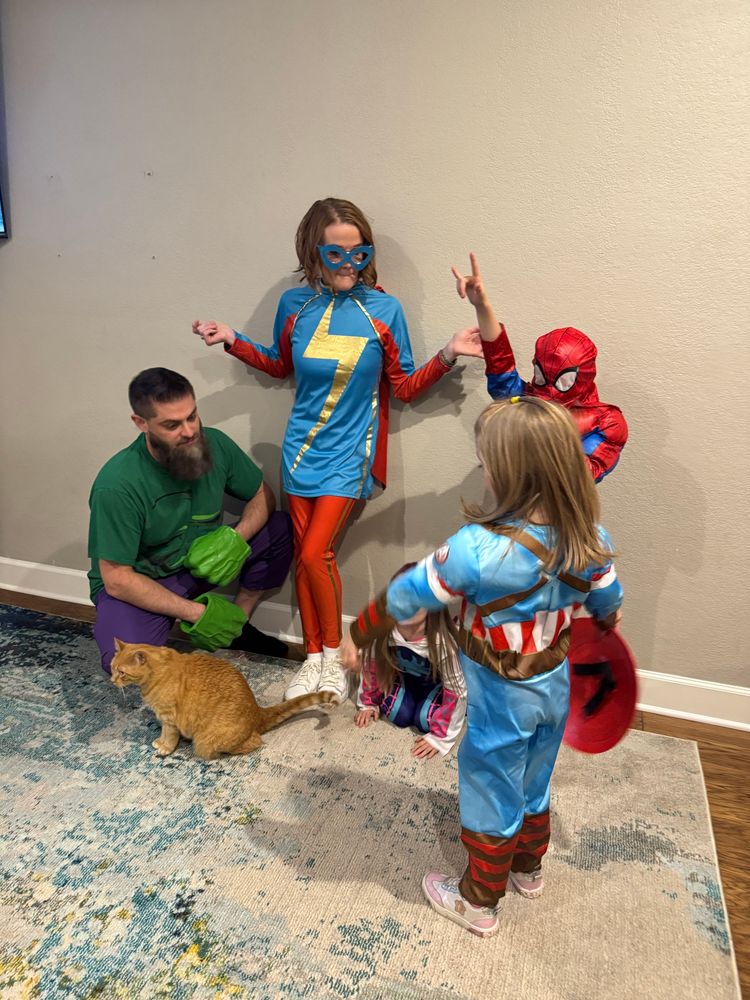 A very unorganized picture of us in Halloween costumes: the Hulk, me as Ms Marvel with both hands in the air like “what are y’all doing?” oldest as Spiderman, middle as Captain America with her back to the camera trying to figure out where to stand, and youngest as Ghost Spider kneeling. Also featuring: Orange cat. 