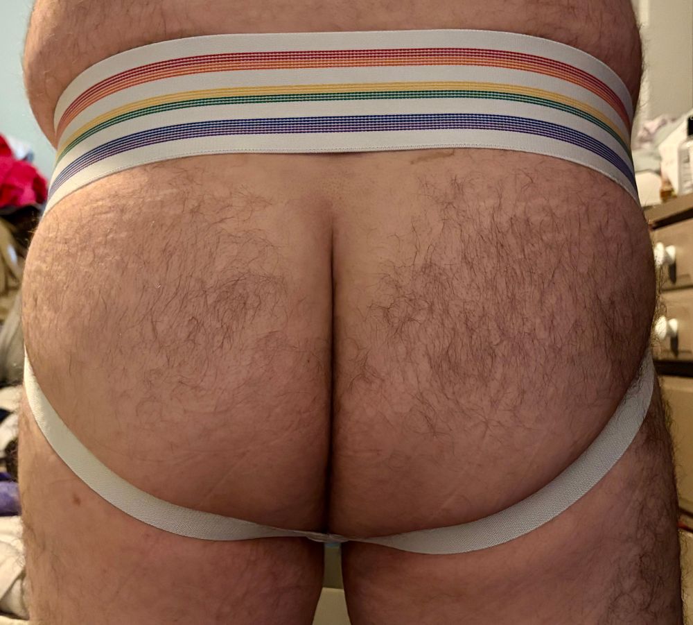 A pic from lower back to upper thighs. A hairy man stands in a white jockstrap with the upper waistband also containing color stripes arranged in rainbow order. The lower straps frame his hairy butt. 