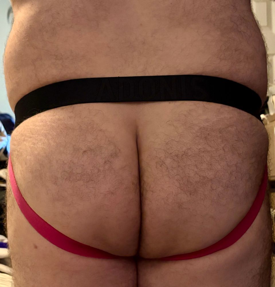 A picture from lower back to upper thighs. A hairy man stands in his jockstrap, showing how it frames his butt. The upper strap is black and the lower straps are pink. 