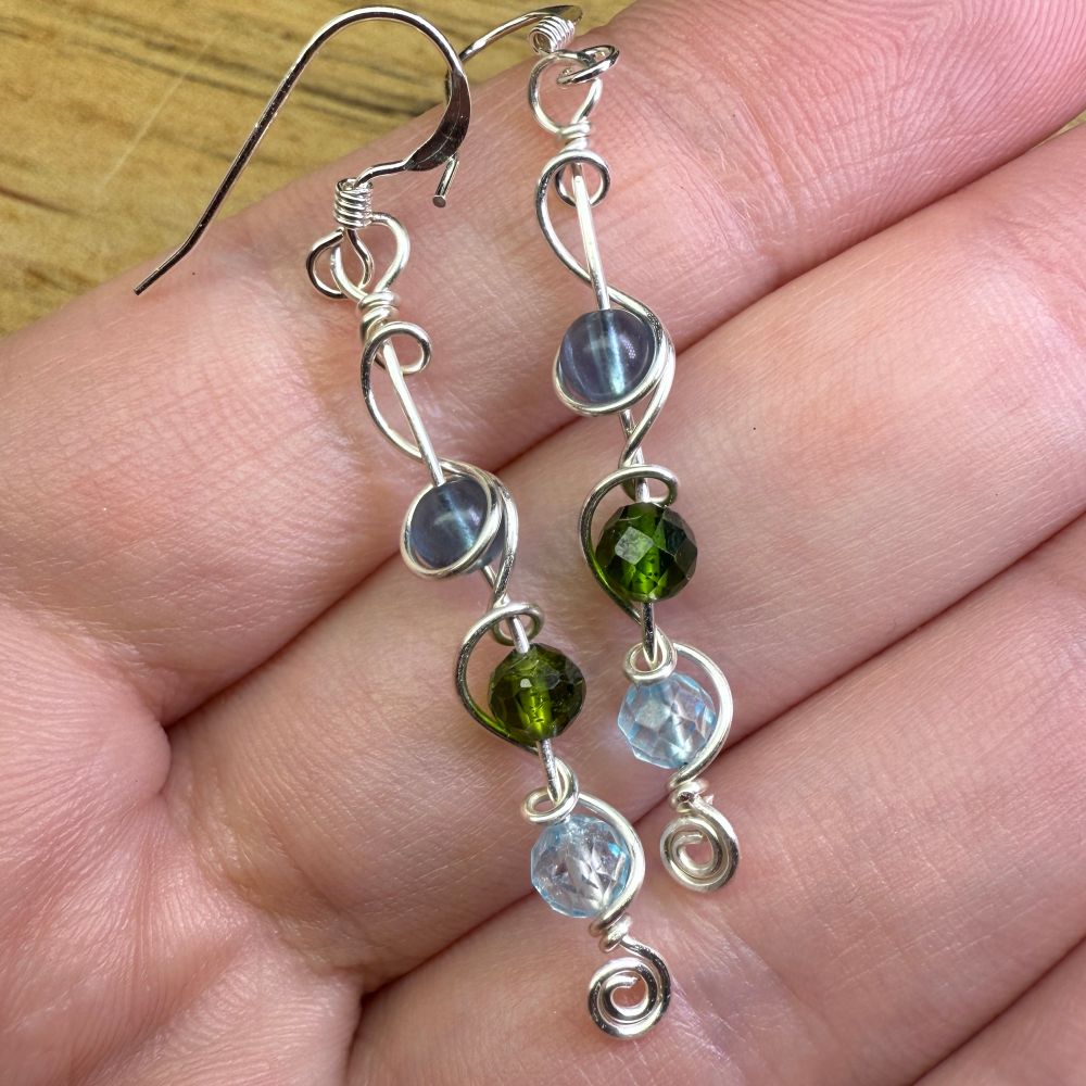 A pair of dangly earrings made from small beads of blue Fluorite, chrome Diopside, and blue Topaz with swirly silver wire, held in a woman’s hand. 
