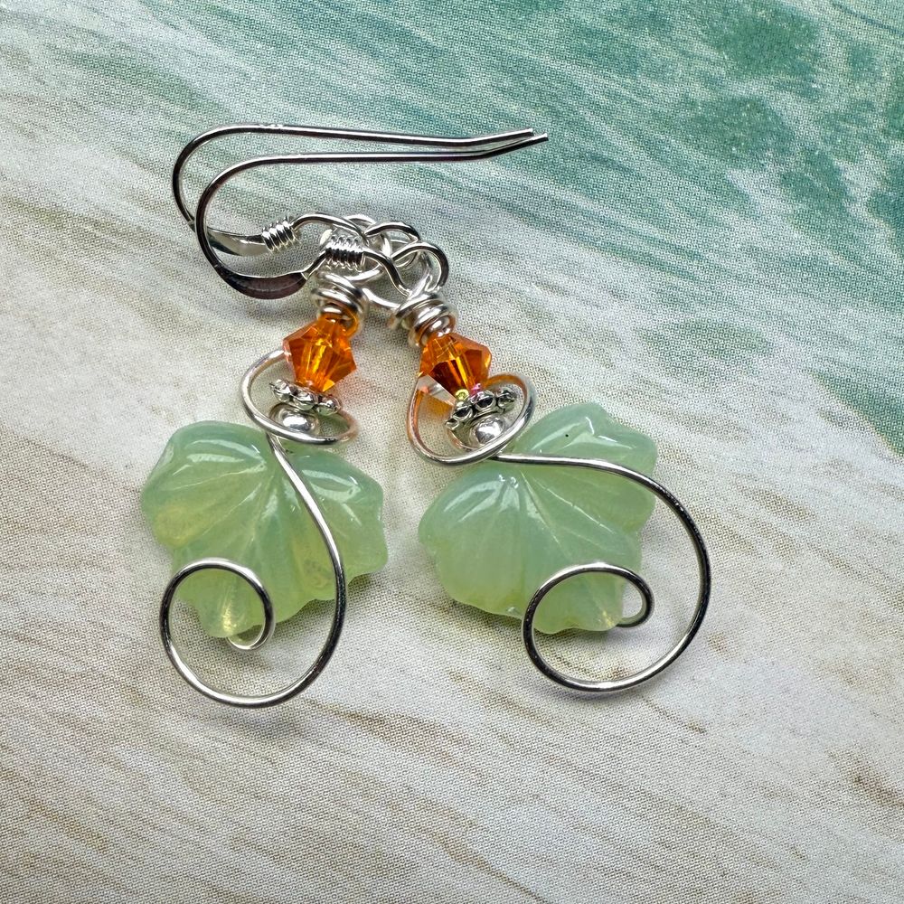 A pair of earrings made from pale green glass leaves and bright orange bicones in swirls of silver wire, resting on a piece of paper that looks like a beach. 