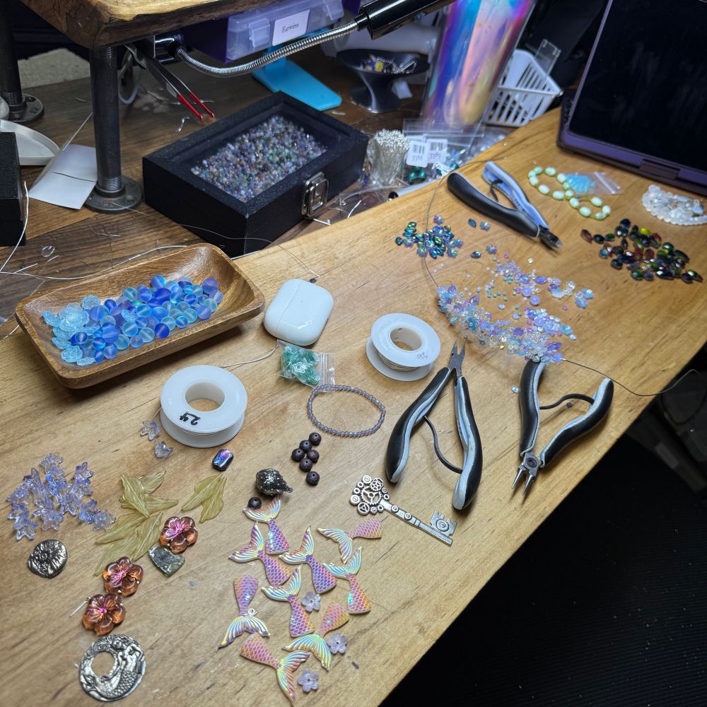 A photo of a long wooden workbench with many piles of beads and other shiny items, plus three pairs of pliers, two small spools of wire, and an AirPods case. There is a partially finished necklace of blue and purple flowers, a pile of iridescent pink mermaid tail charms, a bowl of blue beads, some green kelp-like leaf beads, some pewter charms from Green Girl studios, and a number of other piles of beads that are out of focus. In the background there is a black box with a clear lid filled with gemstones, some small plastic bags with barcode labels on them, N iridescent insulated glass, and an iPad. 