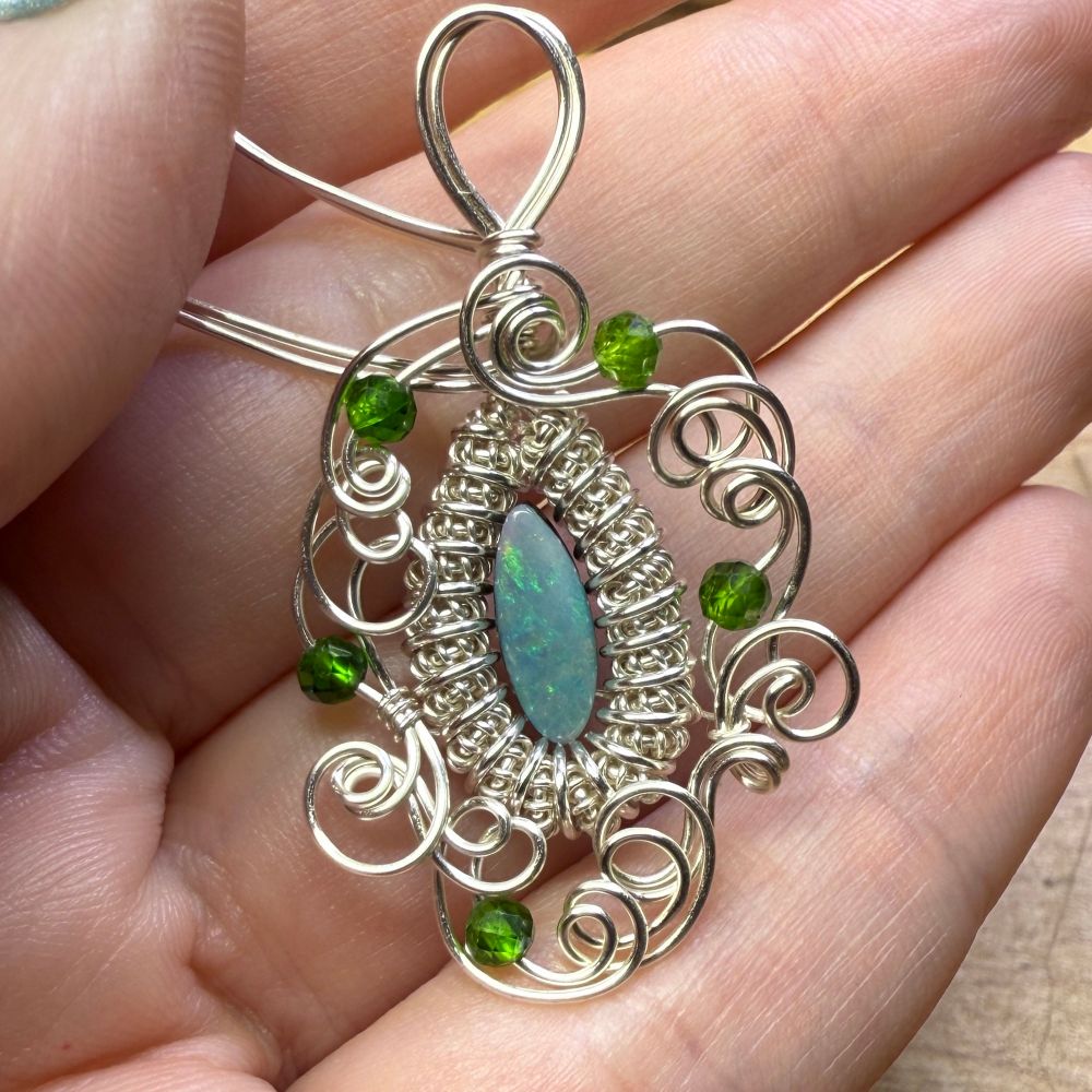A pendant made from an opal doublet with green fire, set in a coiled wire bezel with the start of a swirly frame. There are five very bright green stones placed in the swirly wirework.  