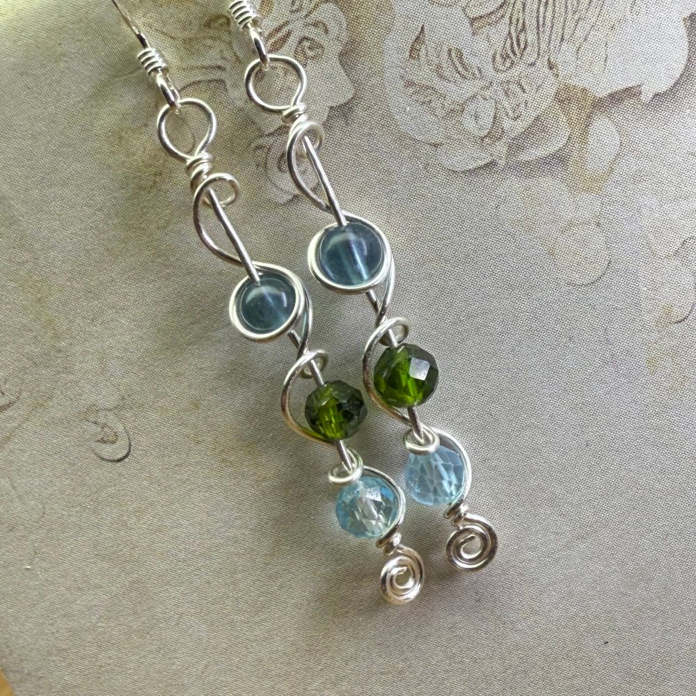A pair of dangly earrings made from small beads of blue Fluorite, chrome Diopside, and blue Topaz with swirly silver wire, photographed against an off-white background. 