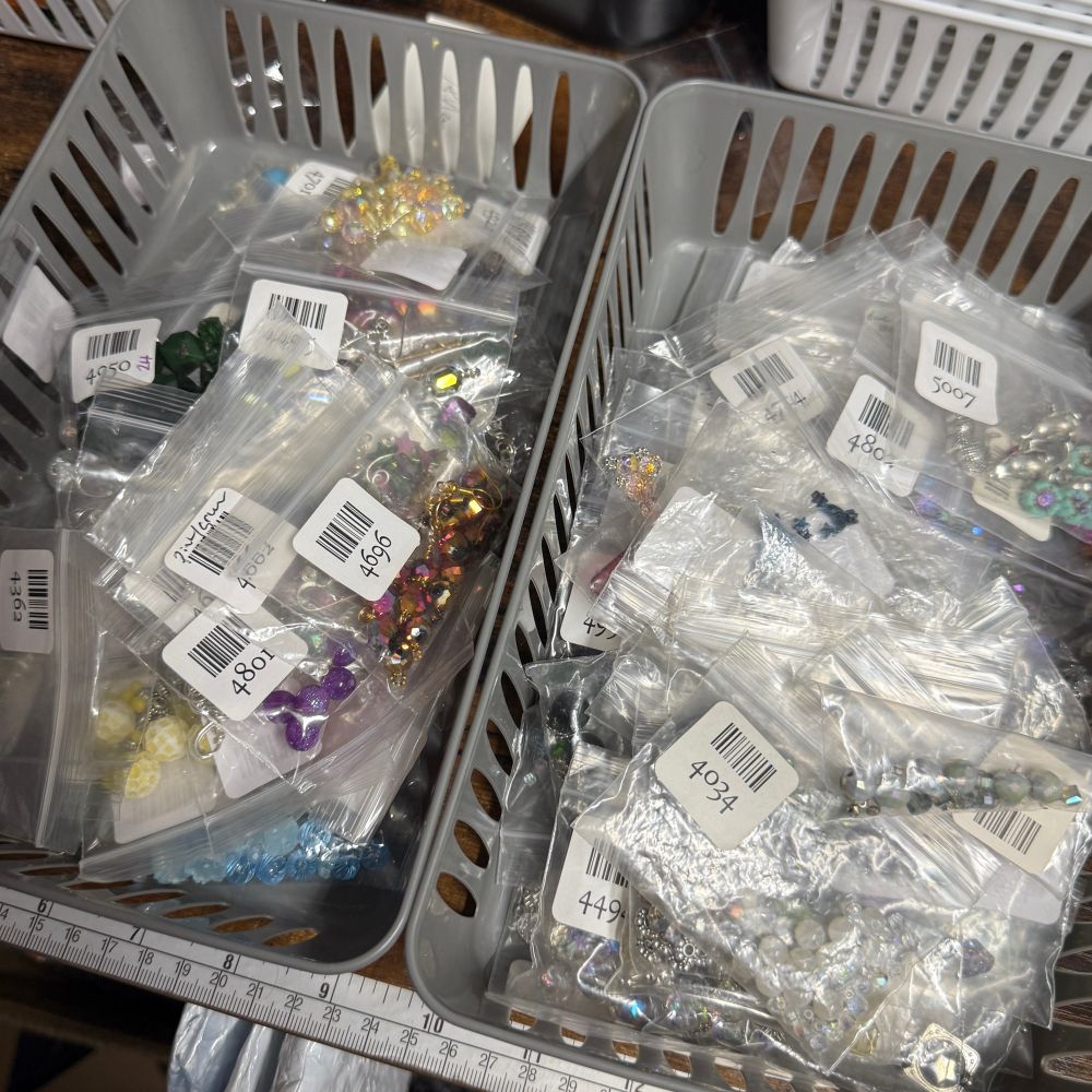 Two grey plastic bins sitting side-by-side on a wooden table. They are filled with small ziplock baggies. Each baggie has a sticker with a barcode and an item number, and colorful beads and pieces of jewelry can be seen in many of the bags. 