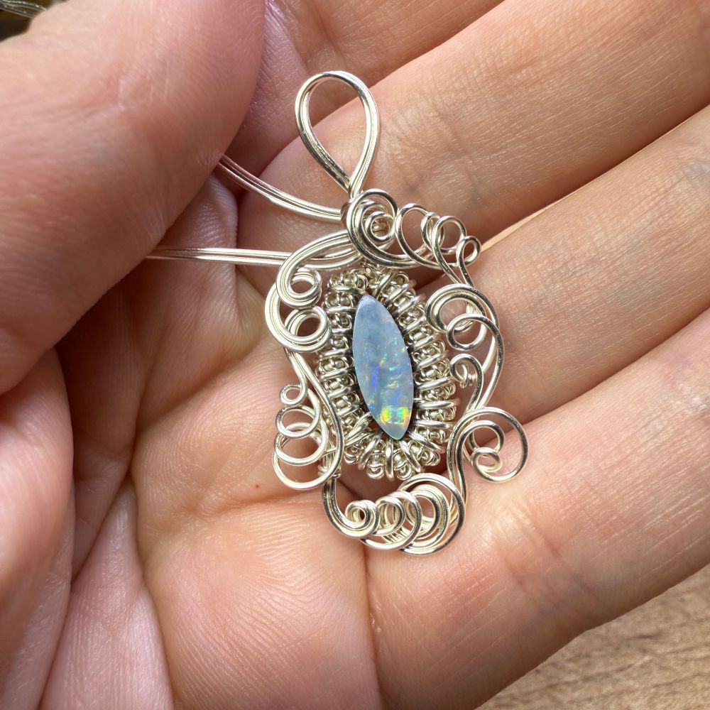A photo of a woman’s hand holding a partially completed opal double pendant. The opal has red and yellow and green and blue side and is set inside a bezel of coiled wire, with a framework of swirly wire outside that. 