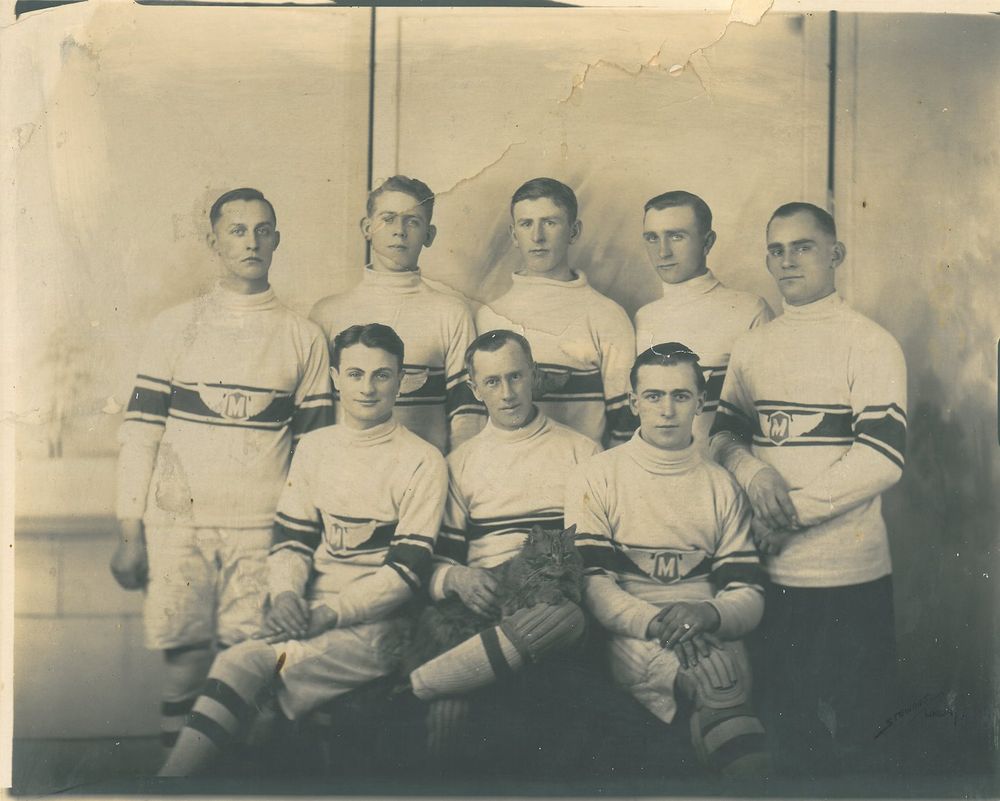 A black and white photo of a hockey team wearing white jerseys with stripes across the middle and the letter M encircled, with wings; circa 1921. One player is holding a cat. 