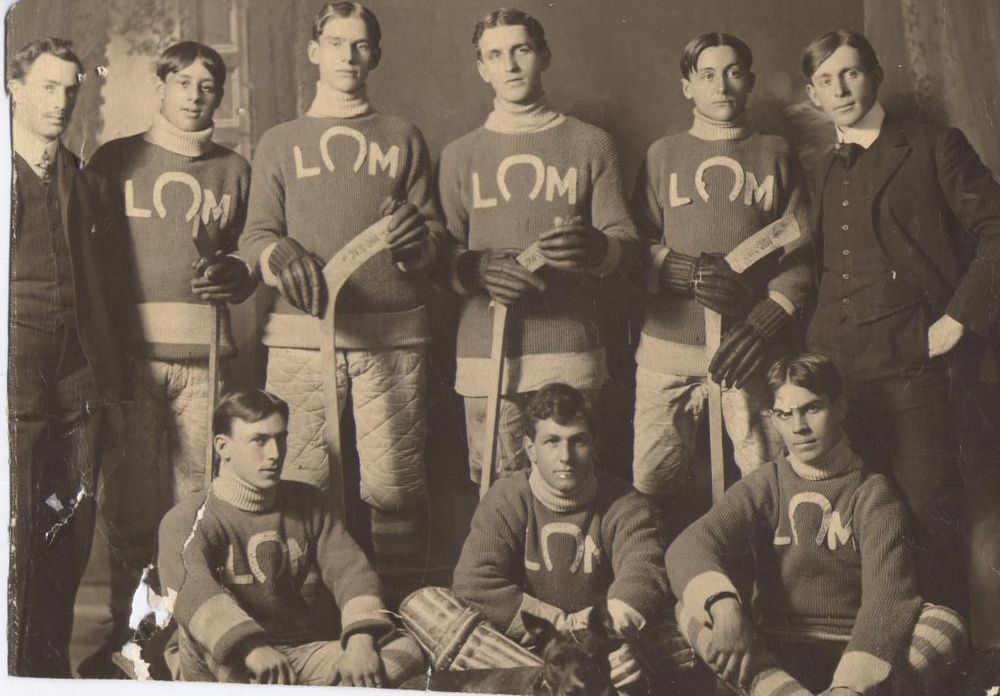 A faded, black and white photo of a hockey team from Lindsay, dating to the early 1900s. Their jerseys feature the letters L and M, separated by a horseshoe or Omega shape. Two men in formal attire stand on either side of the seven players - four players stand in the back row, and three sit in the front row, with the goalie in the middle. 