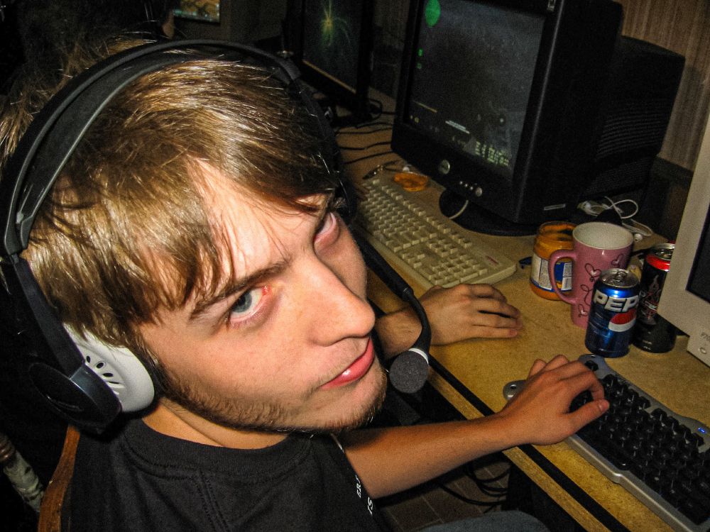 A harsh flash photo of a gamer wearing a headset, glancing over his shoulder at the photographer, with a slight smirk, and some industrial strength stank-eye. A can of pepsi, an energy drink, a jar of queso cheese, and a cute tall pink mug with hearts sits on the desk next to him. several other gaming stations are visible in the background, one of which has a beige Compaq keyboard.