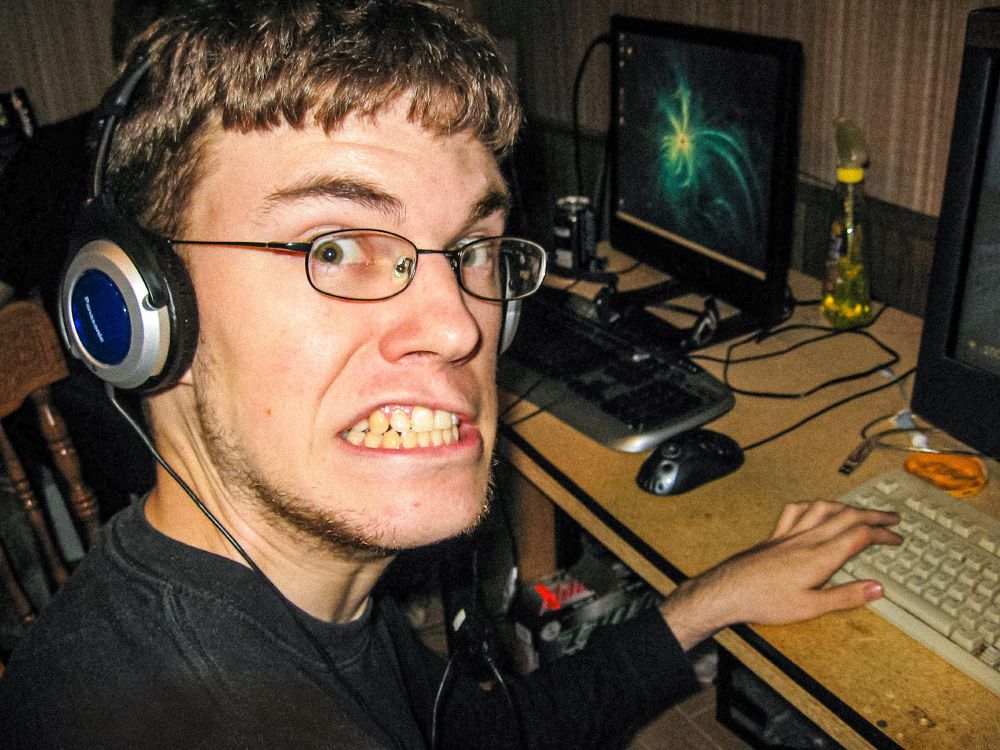 A harsh flash photo of another gamer wearing glasses and a headset, glancing over his shoulder at the photographer, making a very toothy, surprised grimace. They are using a beige keyboard. The lid of a queso cheese jar, and some kind of yellow cleaner spray bottle are on the desk to his left. The next station behind him has a logitech MX518 mouse. A case of some form of Mountain Dew is visible on the floor under the desk. 