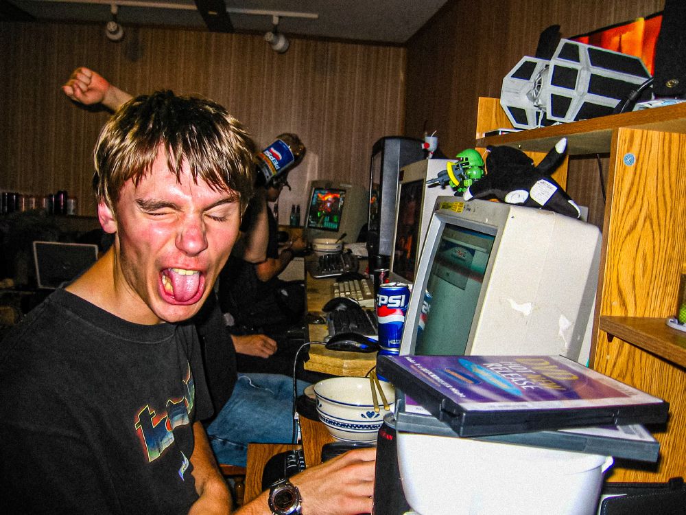 A harsh flash photograph of a short haired nerd wearing a TOOL shirt, facing the camera and squinting hard, and making a "BLEGH" face, while using their computer, with a large CRT monitor. In the foreground are two Blockbuster DVD rentals, sat on top of a coffee machine. In the background are several other players on additional computers with CRT monitors. One obscured player can be seen with a raised fist, and a bottle of pepsi raised in a chugging position. Various cans of cola, energy drinks and bowls previously used for massive amounts of Mr Noodle ramen are strewn about on the desks. Toys of a Tie Fighter (possibly the X1 or X2?), a chibi Zaku, and GIR are visible. A Dell Inspiron 6400 laptop is in the far background. 