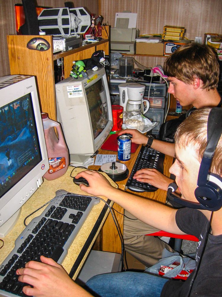 A harsh flash photo of nerds playing what might be WoW on large CRT monitors. A massive jug of chocolate milk with condensation, and a can of Pepsi is visible. A coffee pot is sat at the far end of the desk. the background is cluttered with action figures/statues, blank CD's and various hardware, wires, boxes of things, a can of compressed air, and bulk flats of Mr Noodles. The player in the foreground is wearing a headset with microphone. 