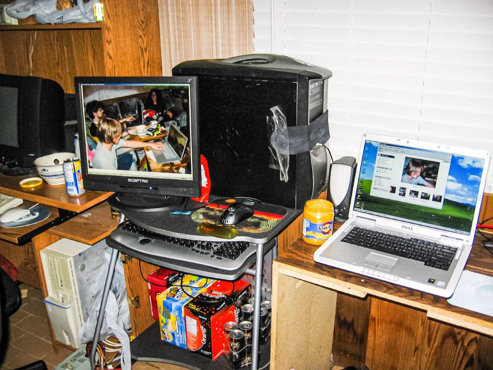 My two computers. The tower PC is a Soyo Dragon case with a handle on top. A black sock is taped to the front of the case, because the LED's on my fan controller/card readers were WAY too bright. Both the PC and the Laptop have other photos from the LAN party on screen. Under the desk is a case of Dr Pepper, a case of Five Alive, a case of Great Value Orange pop, and at least 7 cans of Sobe A-RUSH energy drink. There is a beige IBM Aptiva of sorts in the battlestation next to mine. The Queso cheese makes a glorious return. 