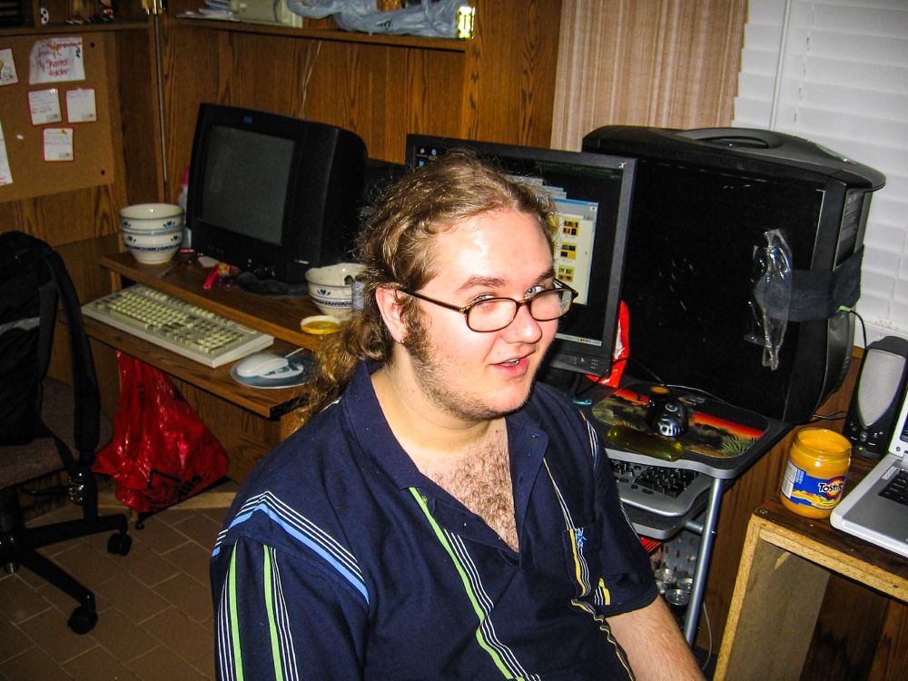 A harsh flash photo of a hairy Jesse, in front of my two computers, a Dell Inspiron 6400, and my Soyo Dragon PC which contained an AMD XP 3000+, and an ATI All-In-Wonder X800. Neither of which made me any better at gaming. An open jar of queso cheese sits next to the laptop. Someone elses battlestation is behind me with a large black CRT, and several empty ramen bowls.  