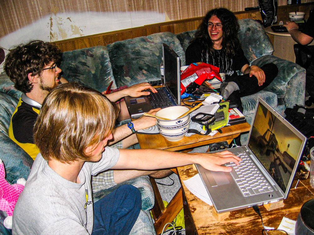A harsh flash photo of 3 guys sitting on a large L shaped sofa, 2 of which are playing CounterStrike: Source. One fellow with medium length blond hair is on a 15 inch 2005 Macbook Pro, and the second with short dark curly hair is on a blue Dell Inspiron 5100 series laptop. The third guy, with long curly hair is at the farthest end of the sofa, laughing and talking with the man on the Dell. Ramen bowls, chip bags, a large MP3 player, and other clutter fills the tables. There is actually a woman only BARELY visible, tucked in the corner of the couch, playing a red Nintendo DS. she is obscured by the Dell fellow.