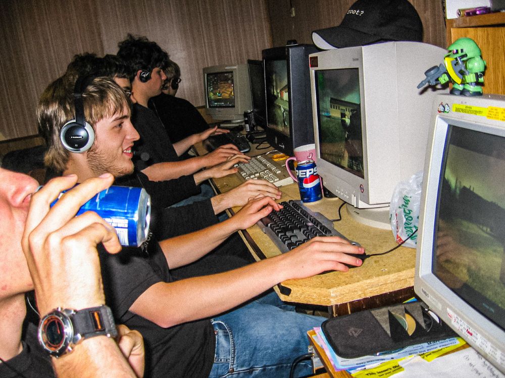 A harsh flash photo of 5 guys in a row, all wearing headsets, playing games at their CRT monitors. The one closest to the camera is drinking a can of pepsi. another can is visible in the background. One player is smiling. On top of his monitor is a ballcap on top that says "Got Root?" on it. 