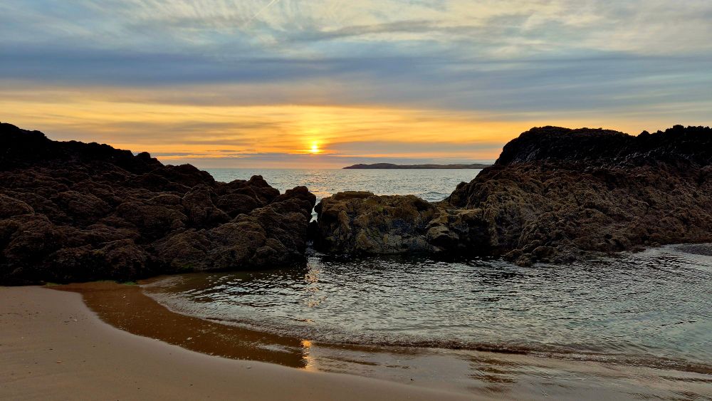 Low orange sunset through streaky pastel clouds and pale blue skies reflected over the sea and rocky beach 