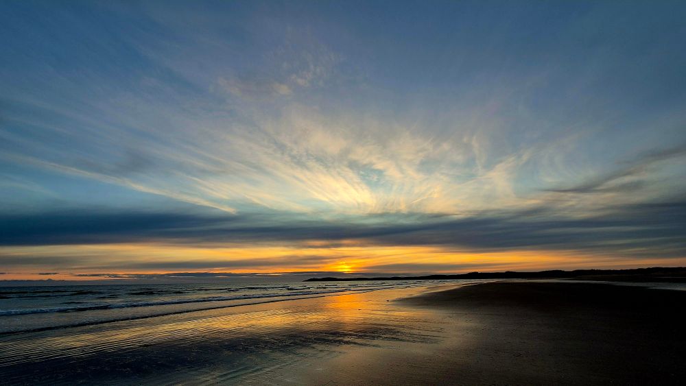 Low orange sunset through streaky pastel clouds and pale blue skies reflected over the sea and wet sandy beach