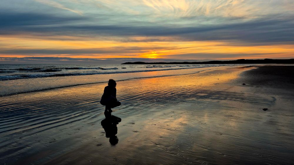 Low orange sunset through streaky pastel clouds and pale blue skies reflected over the sea and wet beach with Liz in the foreground 