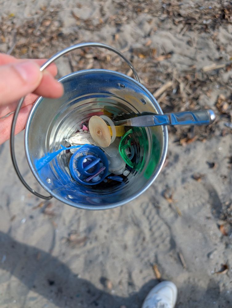 A small metal bucket containing colourful pieces of plastic debris. Sand beach in the background. 